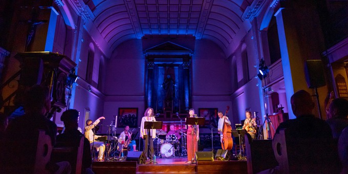 Arun Ghosh’s The Canticle of the Sun, Spitalfields Music Festival, 2022. A band plays on stage with a purple backdrop of the church, with audience members sitting in pews on the foreground. Photo by James Berry.