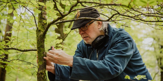 A photo of artist Jez riley French standing in the Forest of Dean. He is a white man, wearing glasses and a flat cap, and wearing headphones, listening to sounds he is recording in the forest. He is surrounded by trees.