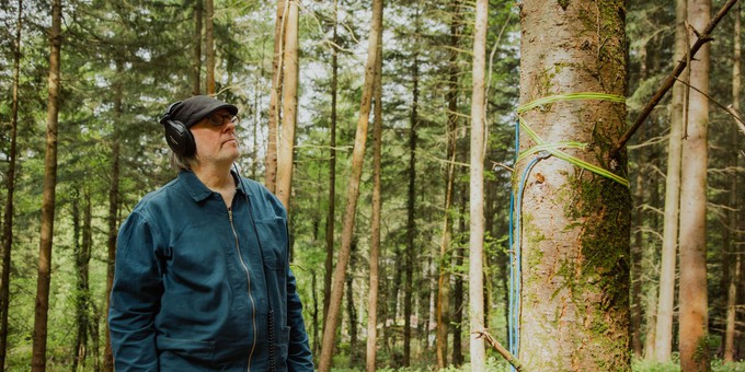 A photo of artist Jez riley French standing in the Forest of Dean. He is a white man, wearing glasses and a flat cap, and wearing headphones, listening to sounds he is recording in the forest. He is surrounded by trees.