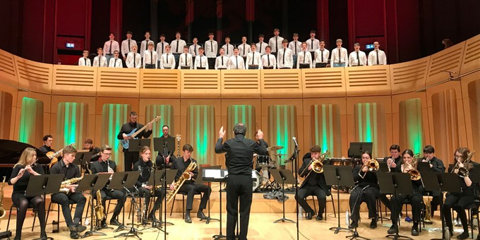A Song for Wales being performed at Royal Welsh College of Music & Drama, March 2022. Royal Welsh College of Music and Drama Big Band are in the foreground, wearing black clothing and playing a range of instruments. The conductor stands in the centre with his arms raised. Above them on the balcony stand two rows of choir members from Only Boys Aloud.