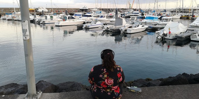 Image of Nikki Sheth wearing headphones and looking out at a harbour full of sailing boats.