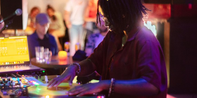 A photo of NikNak performing Sankofa at Strange Brew in Bristol. NikNak is standing at her decks (turntables), with both hands on the vinyl. She is wearing a purple shirt and has bracelets on both arms. Out of focus, there are audience members in the background listening to the music. Photo by Chris Lucas.