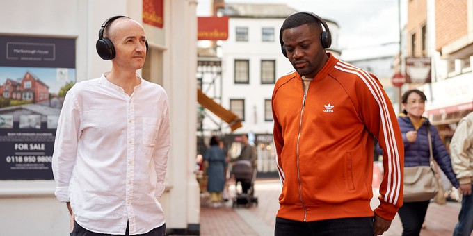Richard Bentley stands on the left. He is a white male, wearing a white shirt and black headphones. He's looking into the distance. Aundre Goddard is standing on the right. He is a black male, wearing an orange top with white stripes on the arms. He is also wearing black headphones and is looking towards the floor. They are standing on Reading High Street. To the right of the picture you can see a female shopper walking past, with black hair, wearing a blue top & wearing a grey handbag. Photo by James John.