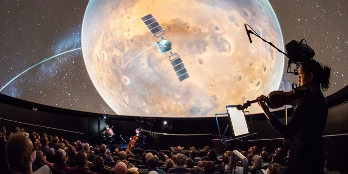The Planets 2018 A photo from The Planets 2018 - a vioinist stands playing as a seated audience watch a satelite in front of the moon on a large cinema screen.