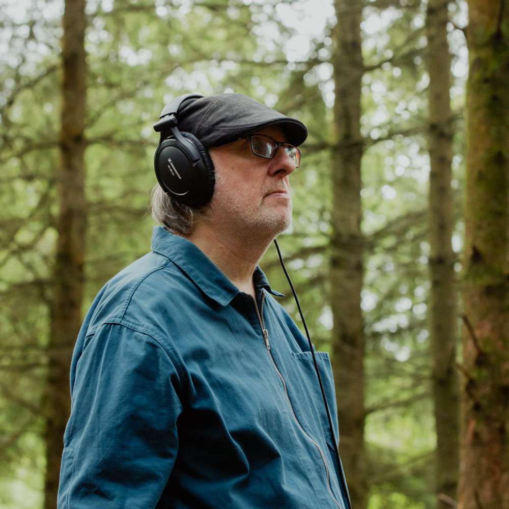 A photo of artist Jez riley French standing in the Forest of Dean. He is a white man, wearing glasses and a flat cap, and wearing headphones, listening to sounds he is recording in the forest.